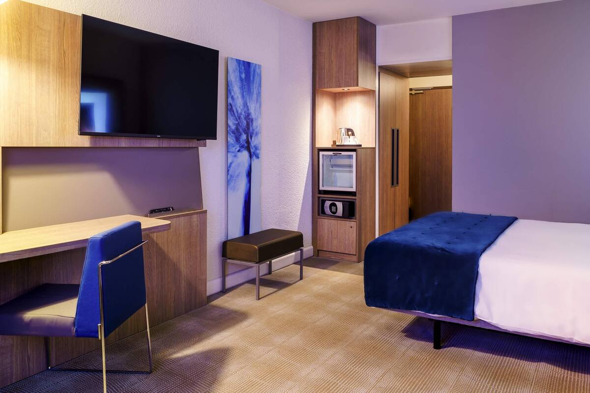A hotel room with a bed, a desk, a chair, a television, a microwave, and a minibar.