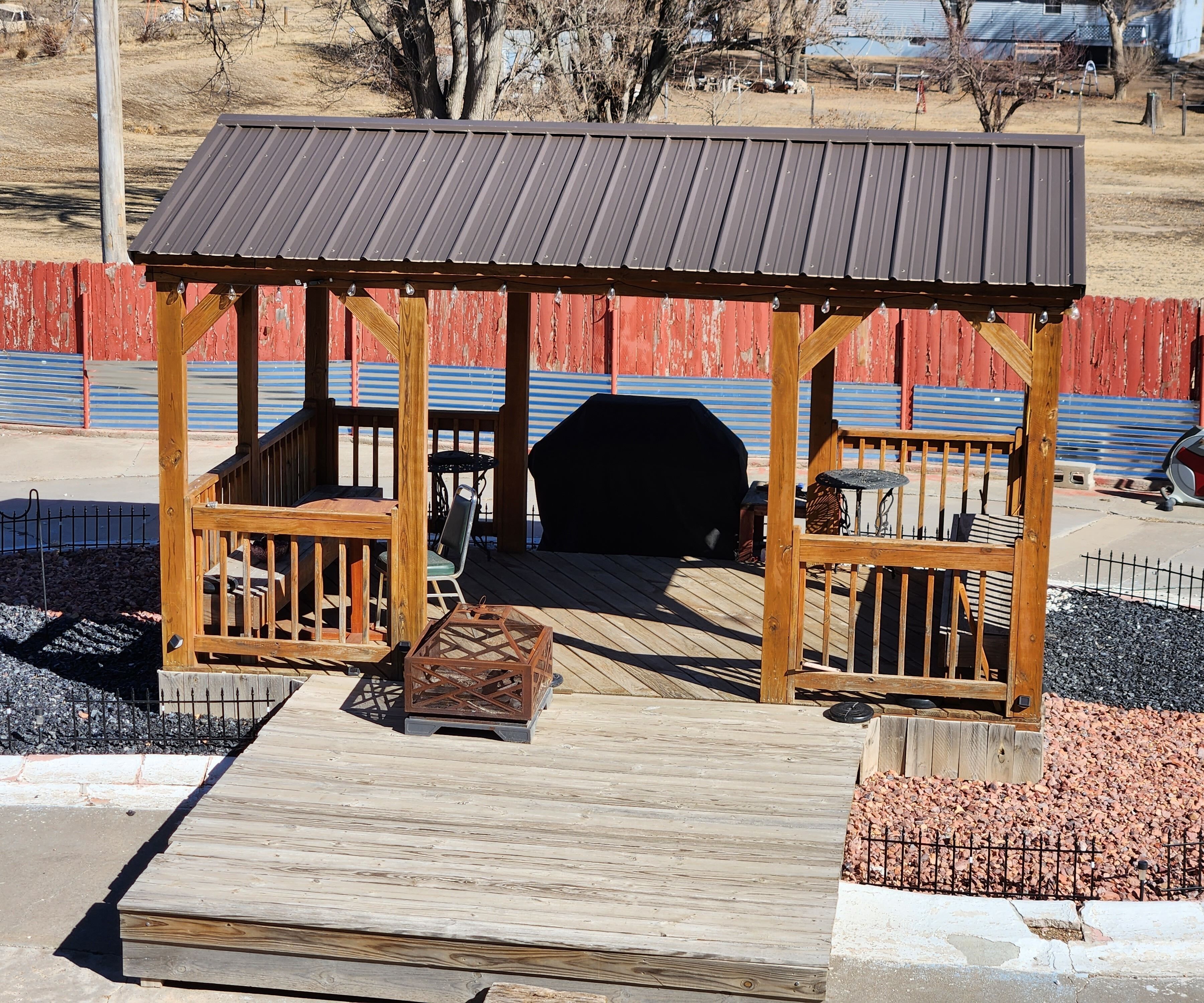 A wooden gazebo with a metal roof, surrounded by a wooden deck and gravel.