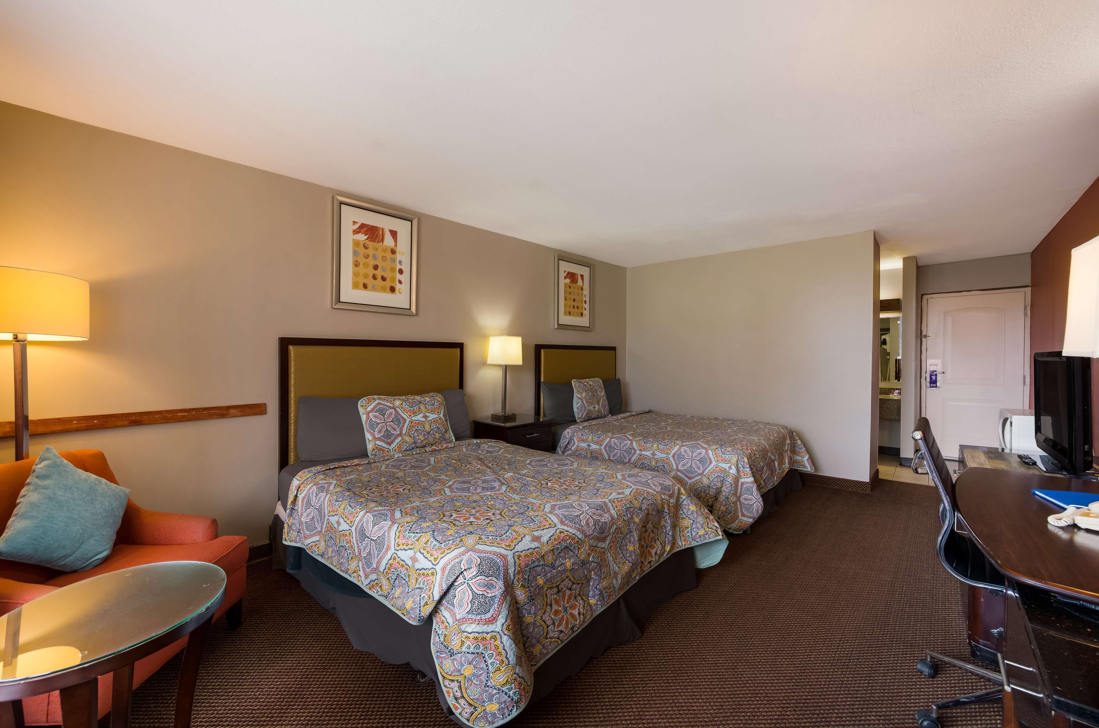 Habitación de hotel con dos camas, un escritorio, una silla, una lámpara y cuadros en las paredes.