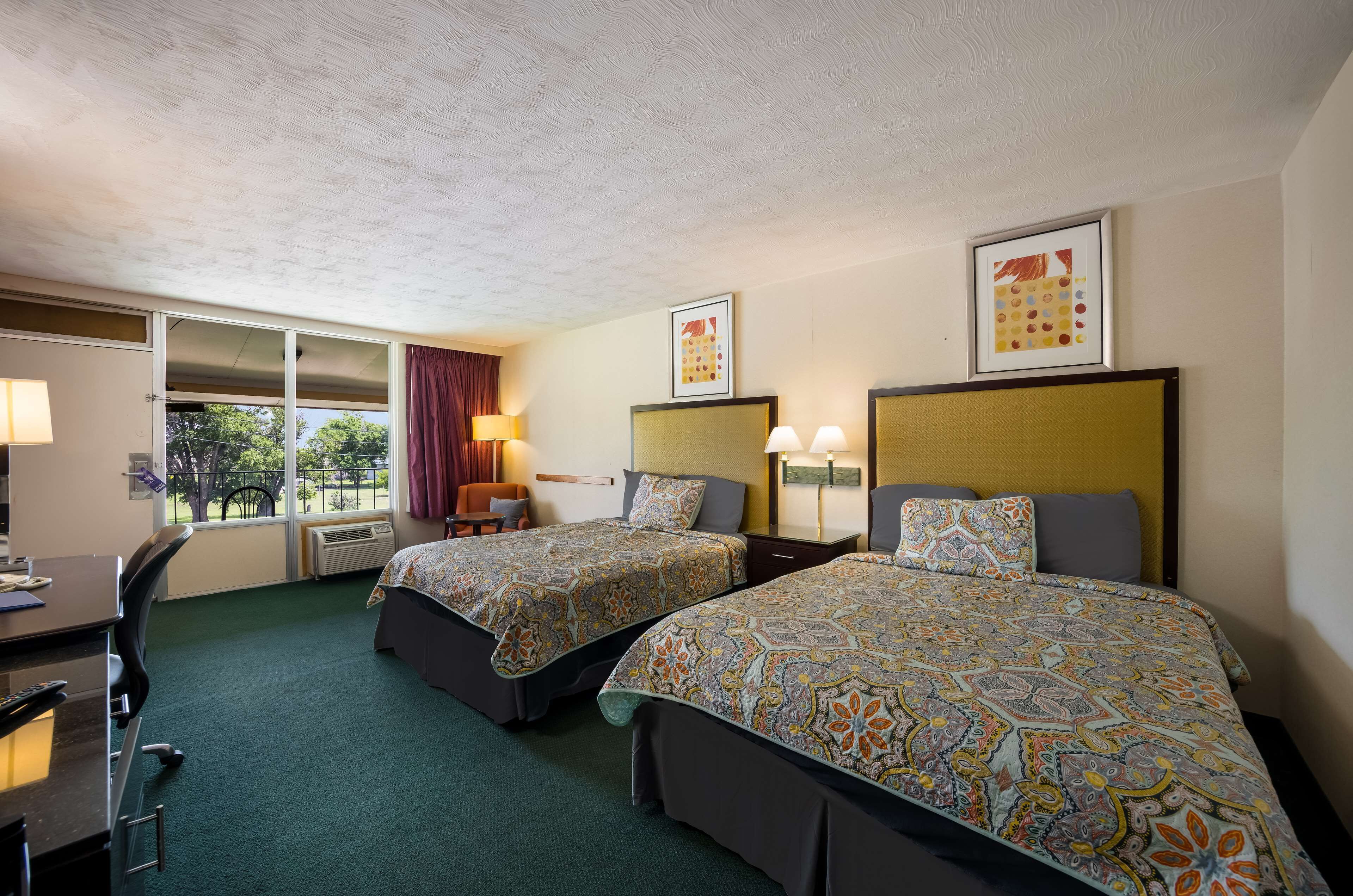 A hotel room with two beds, a desk, and a chair. There are two framed pictures on the wall, a window with curtains, and an air conditioning unit.