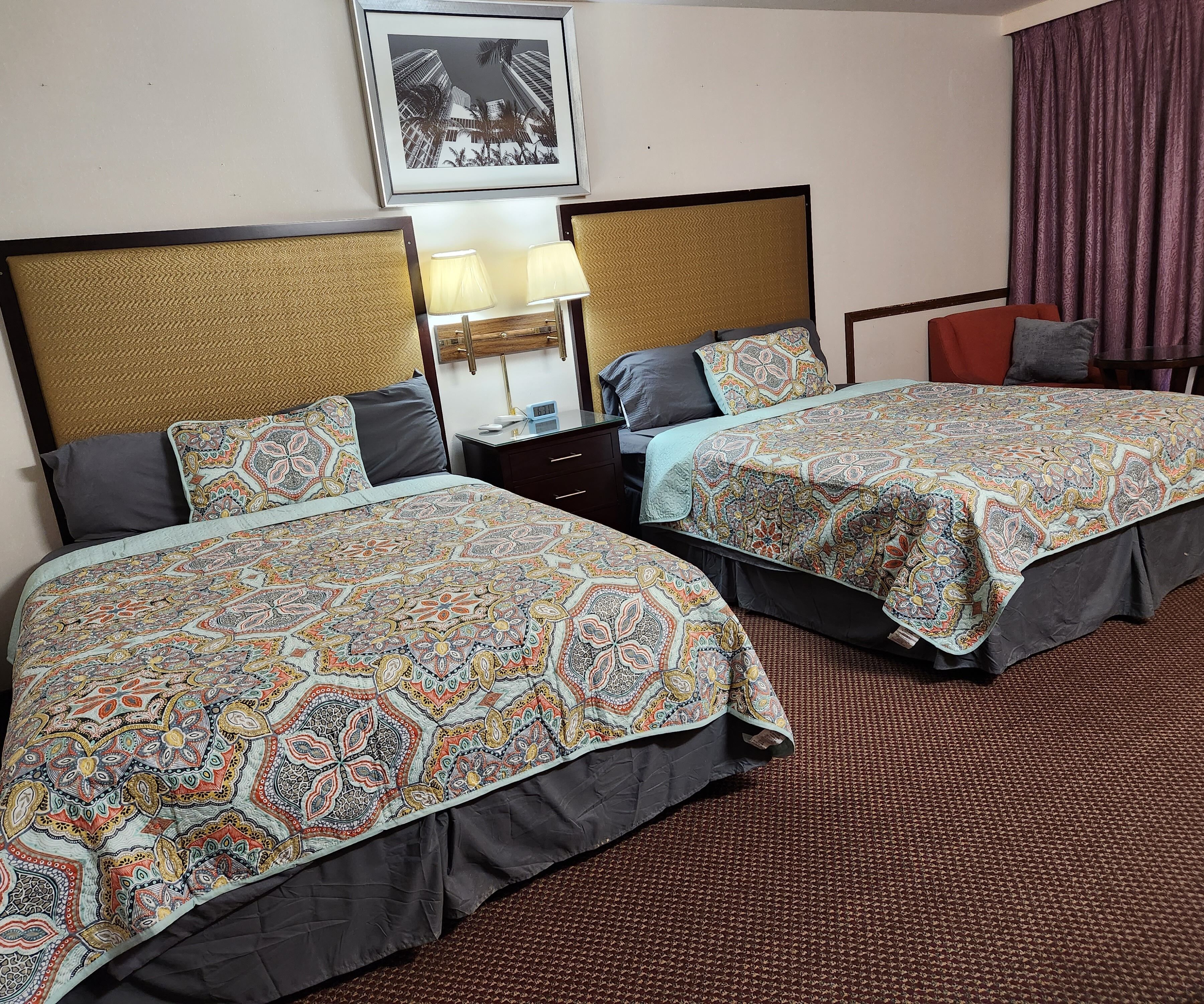 Habitación de hotel con dos camas, una mesita de noche, una lámpara, un cuadro enmarcado en la pared y una ventana con cortinas.