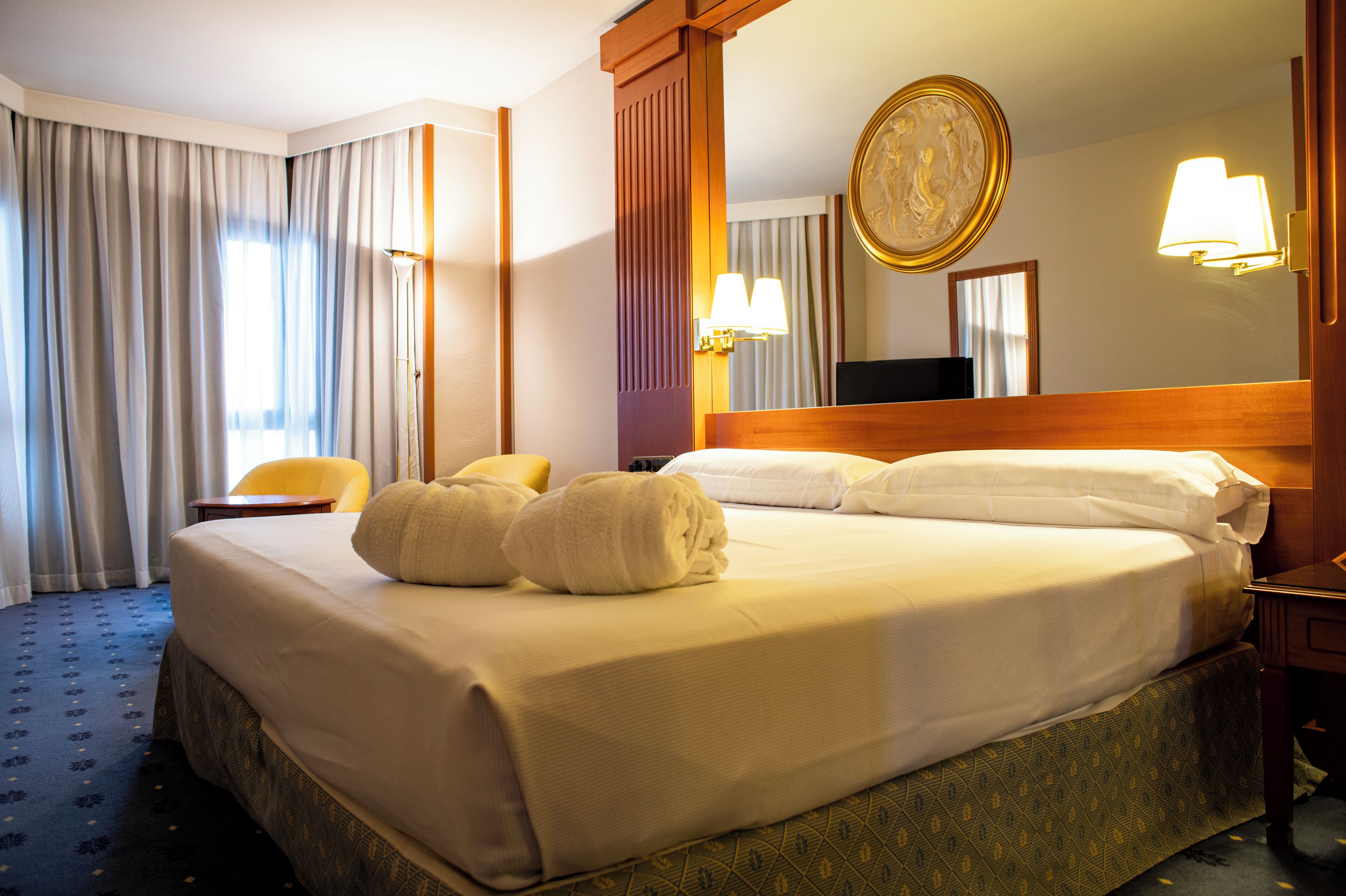 A hotel room with a large bed, two pillows, a bedside table, a chair, a lamp, and a mirror.