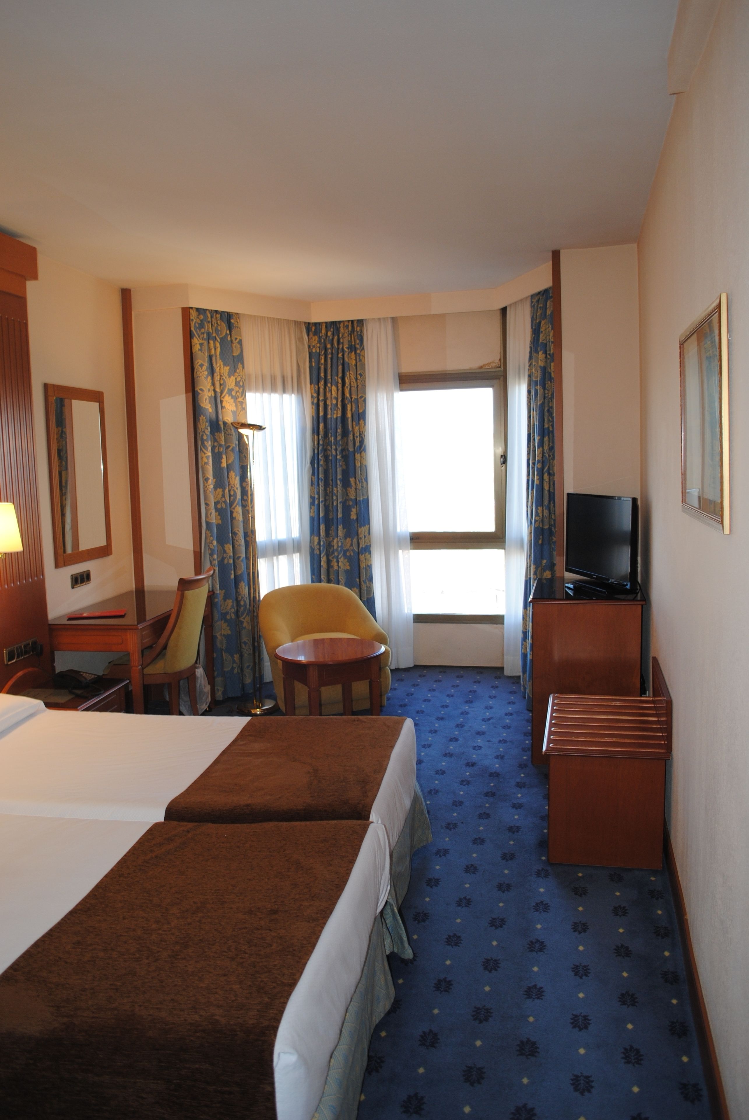 A hotel room with two beds, a desk, a chair, a television, and a large window with curtains.