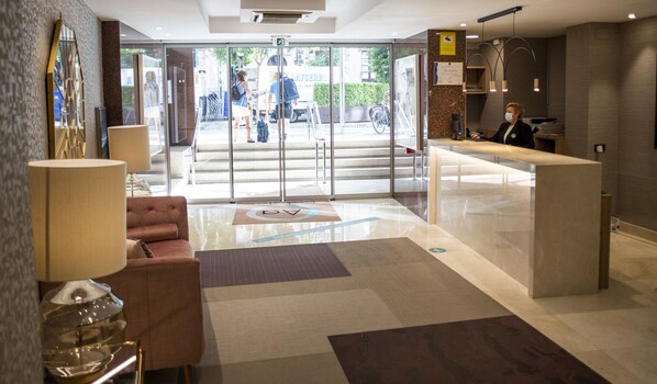 A hotel reception area with a modern design, including a reception desk, a seating area with a sofa, and a glass door entrance leading outside.