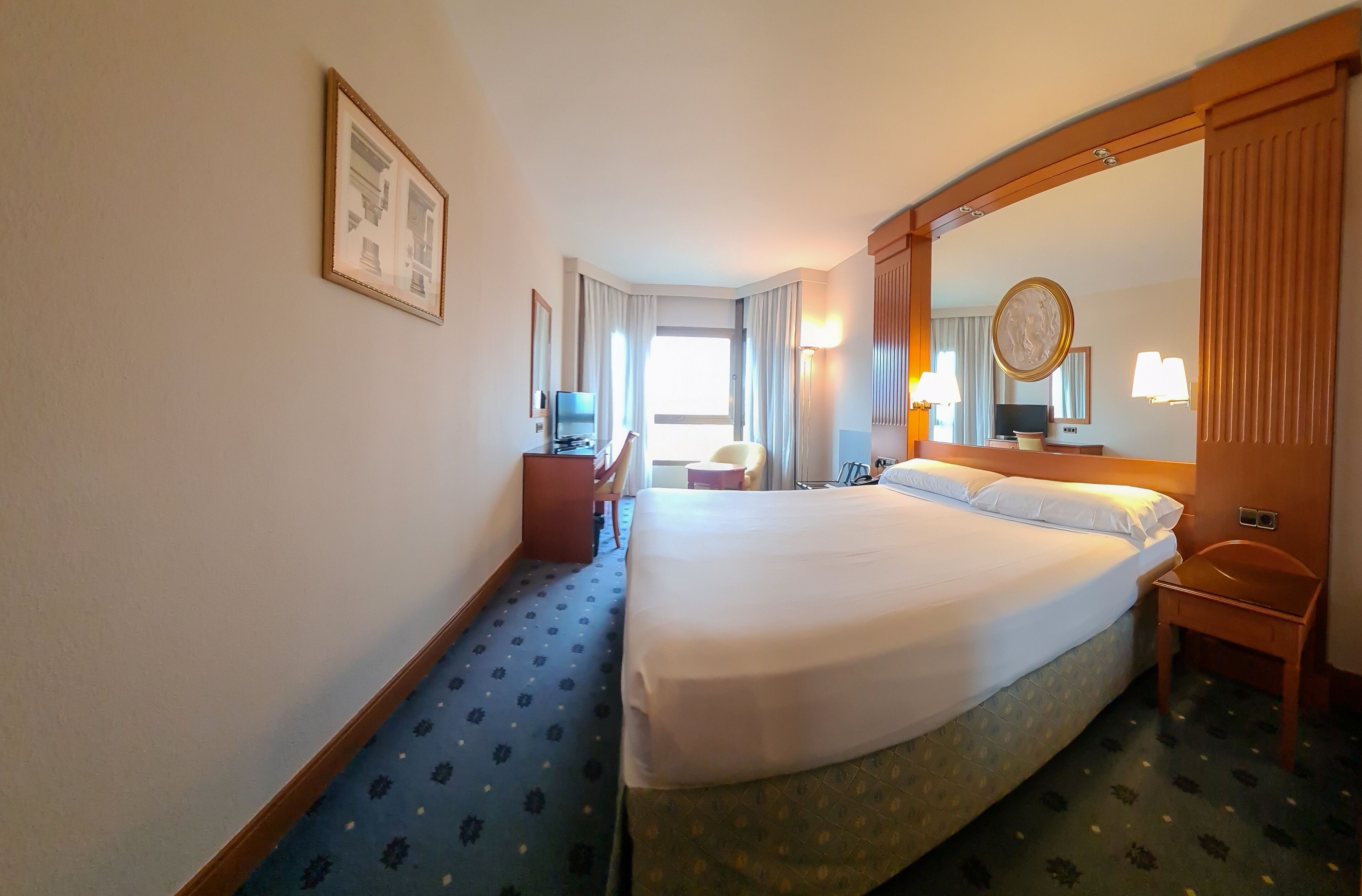 A hotel room with a large bed, a desk with a chair, a television, and a mirror.