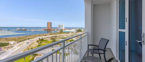 A balcony with two chairs, a view of a road, and a body of water with buildings in the distance.