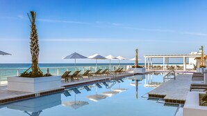Uma área à beira da piscina com espreguiçadeiras e guarda-sóis, vista desobstruída para o mar e piscina refletindo o ambiente.