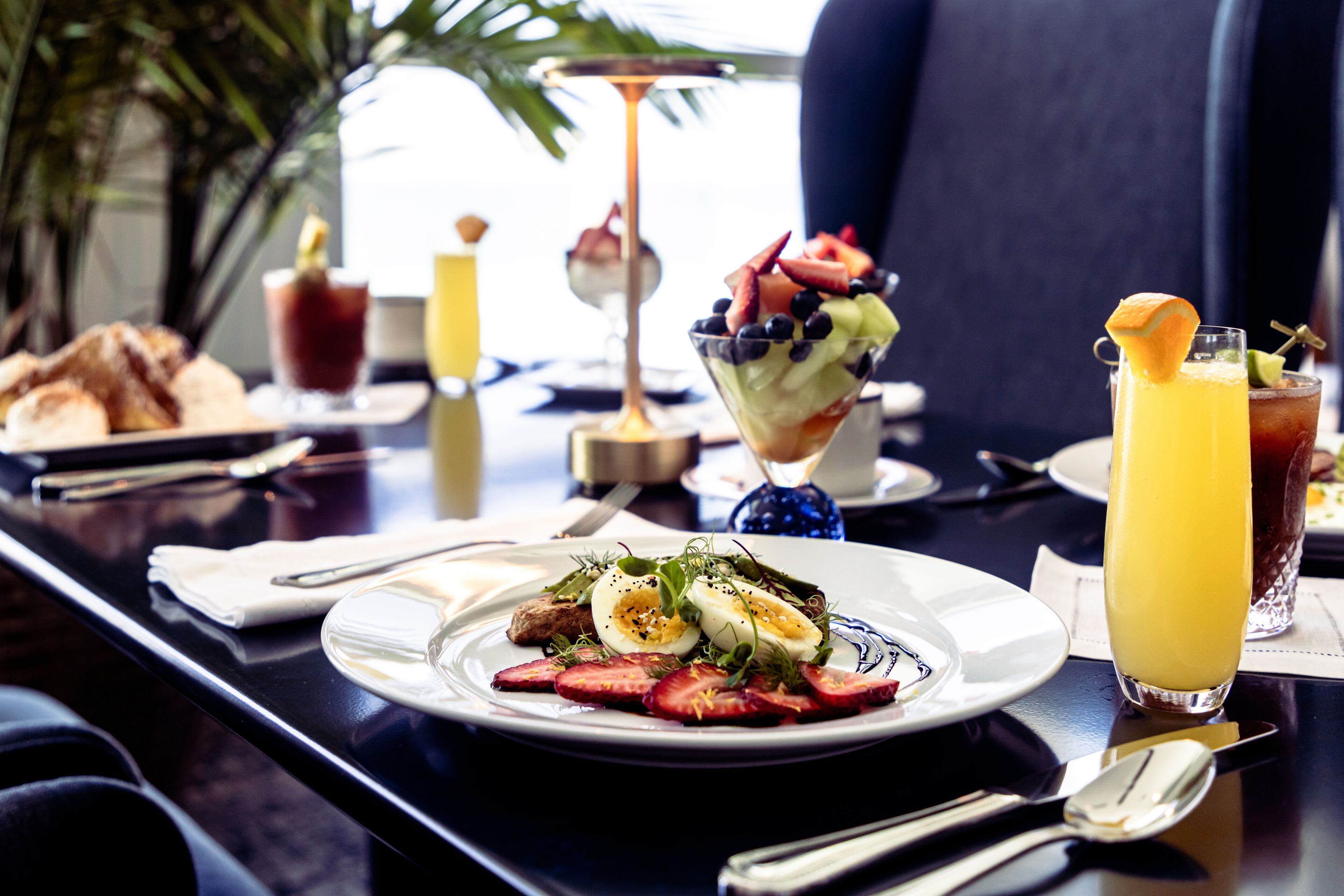 A breakfast setting with a plate of eggs, strawberries, and greens, a glass of orange juice, and a dessert with berries and cream.