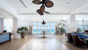 Um hall amplo com balcão de recepção, grandes vasos de plantas, uma mesa central e áreas de estar.