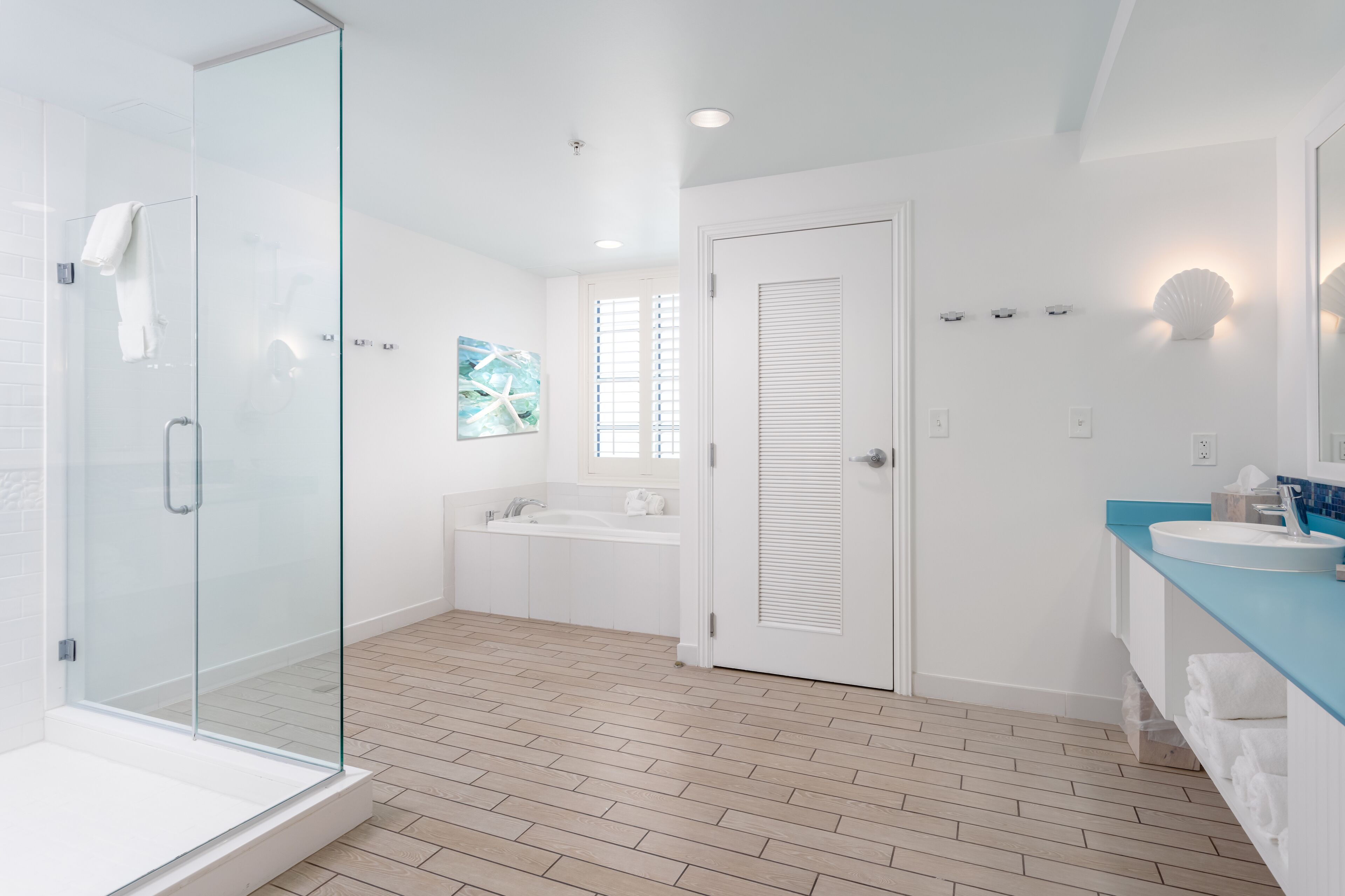 A modern bathroom with a glass shower enclosure, a bathtub, a sink with a blue countertop, and a door with louvered panels.