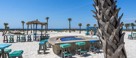A beachside bar area with wooden tables and benches, a central fire pit, and palm trees.