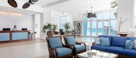 A spacious lobby with a reception desk, seating area with blue upholstered chairs and sofas, a glass coffee table, and a large window overlooking the ocean.