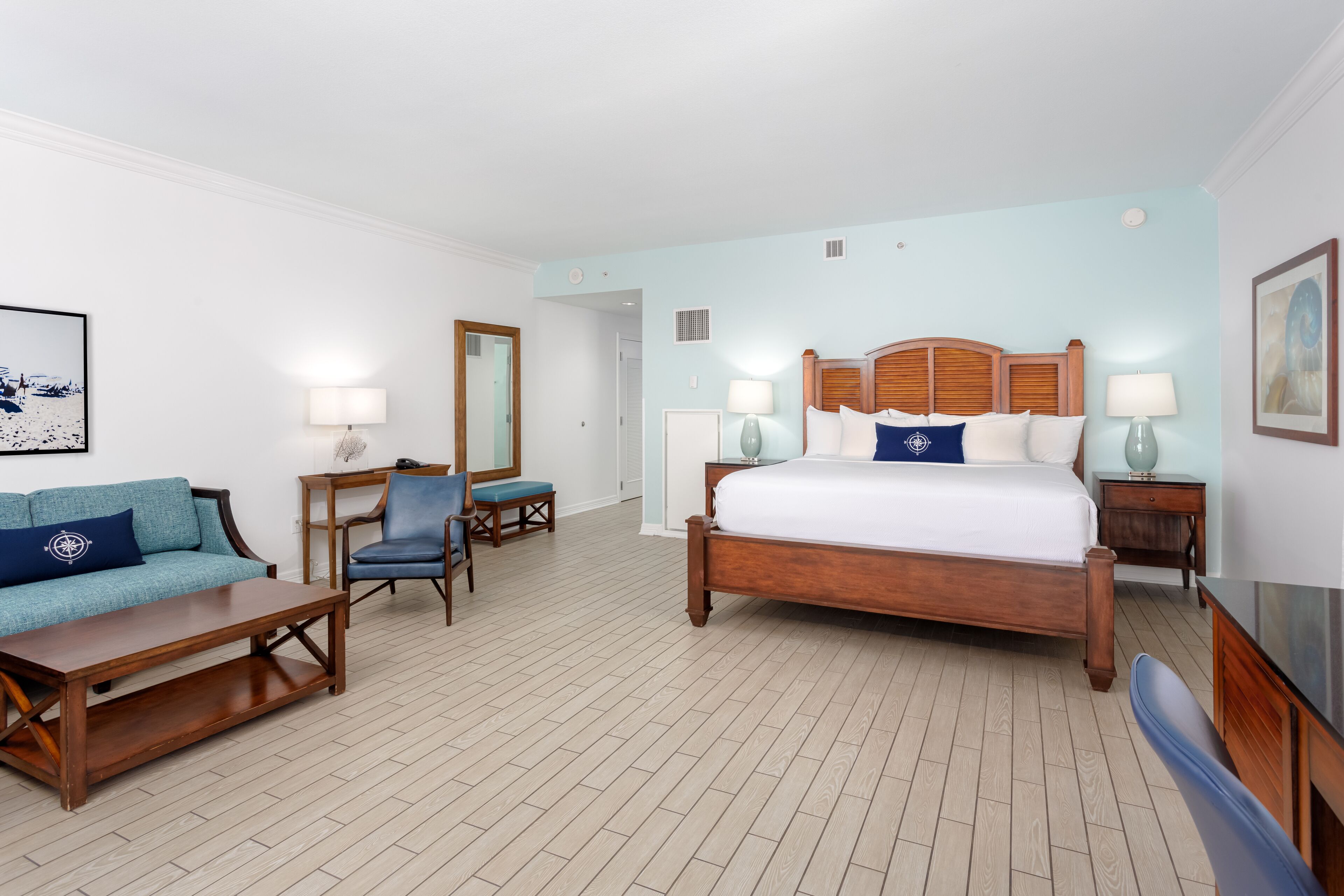 A hotel room with a wooden bed, a blue sofa, a wooden coffee table, a chair, and a desk with a lamp.