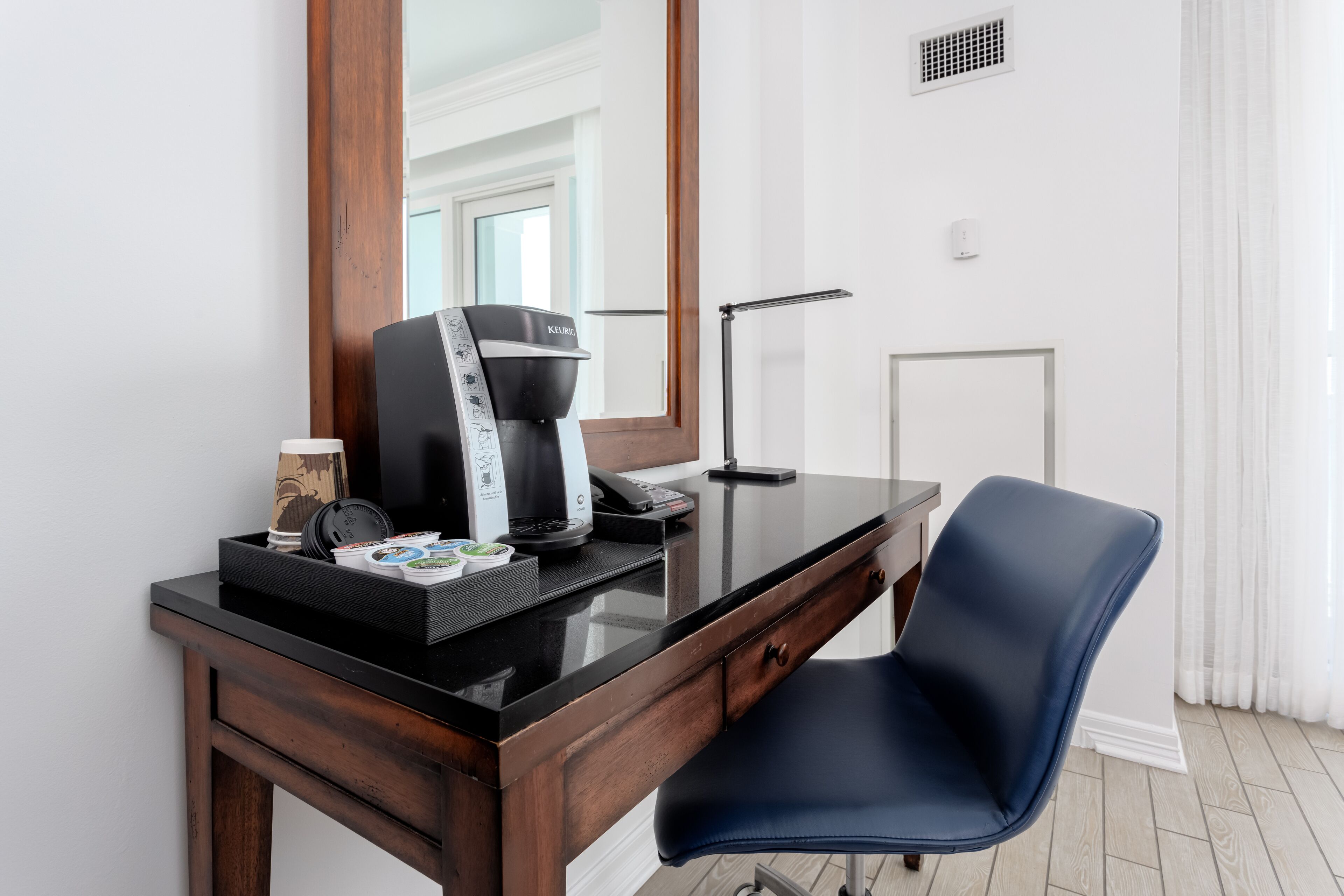 Une chambre d’hôtel moderne avec un bureau en bois, une chaise bleue, une machine à café et un grand miroir.