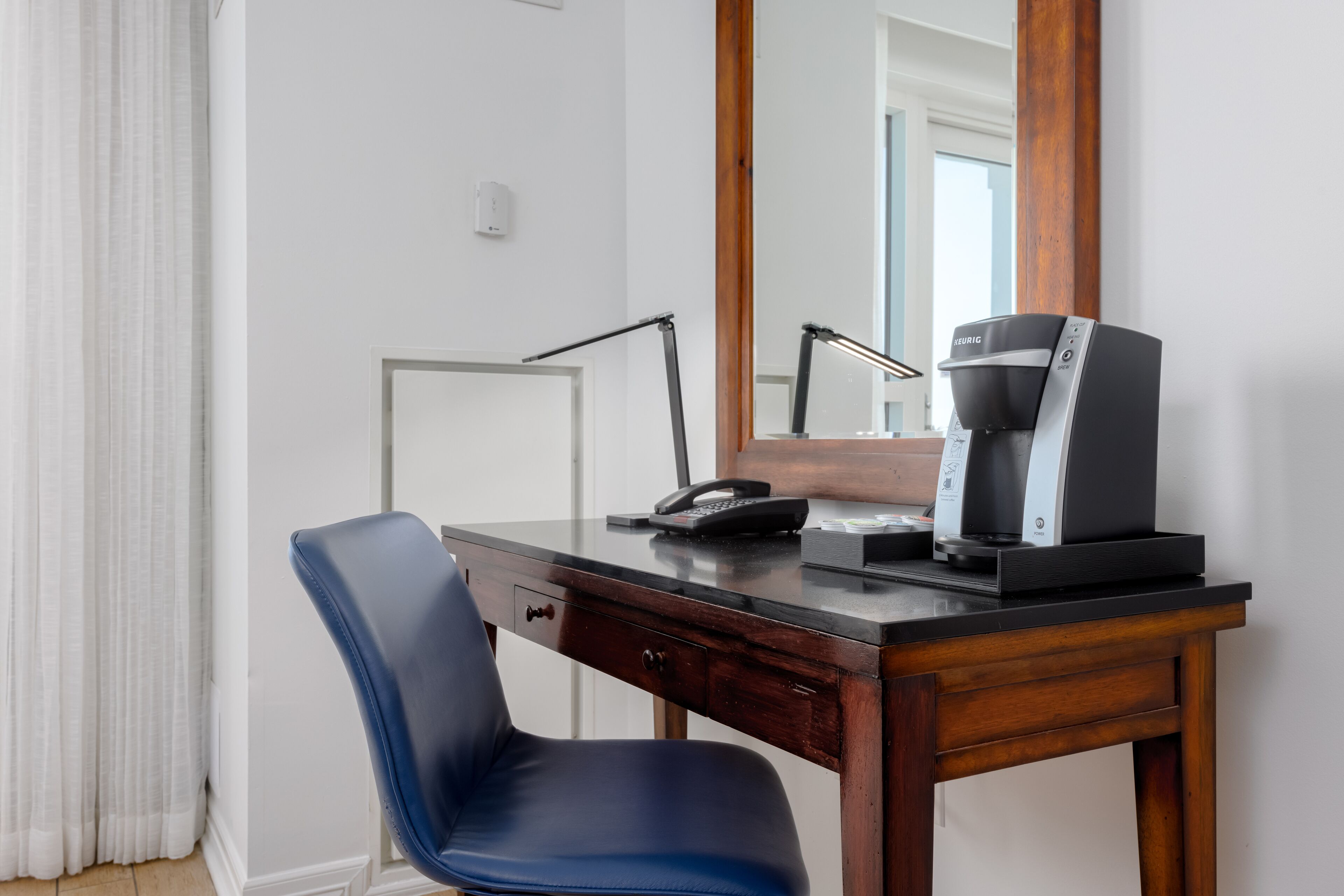 A hotel room with a wooden desk, a blue chair, a coffee maker, and a large mirror.