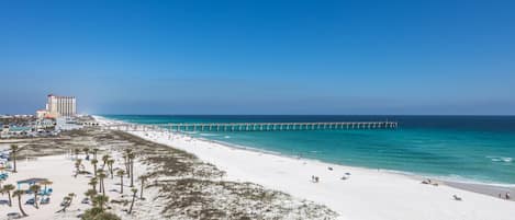 En strand med hvidt sand, en mole der strækker sig ud i havet, og en bygning med rødt tag i baggrunden.