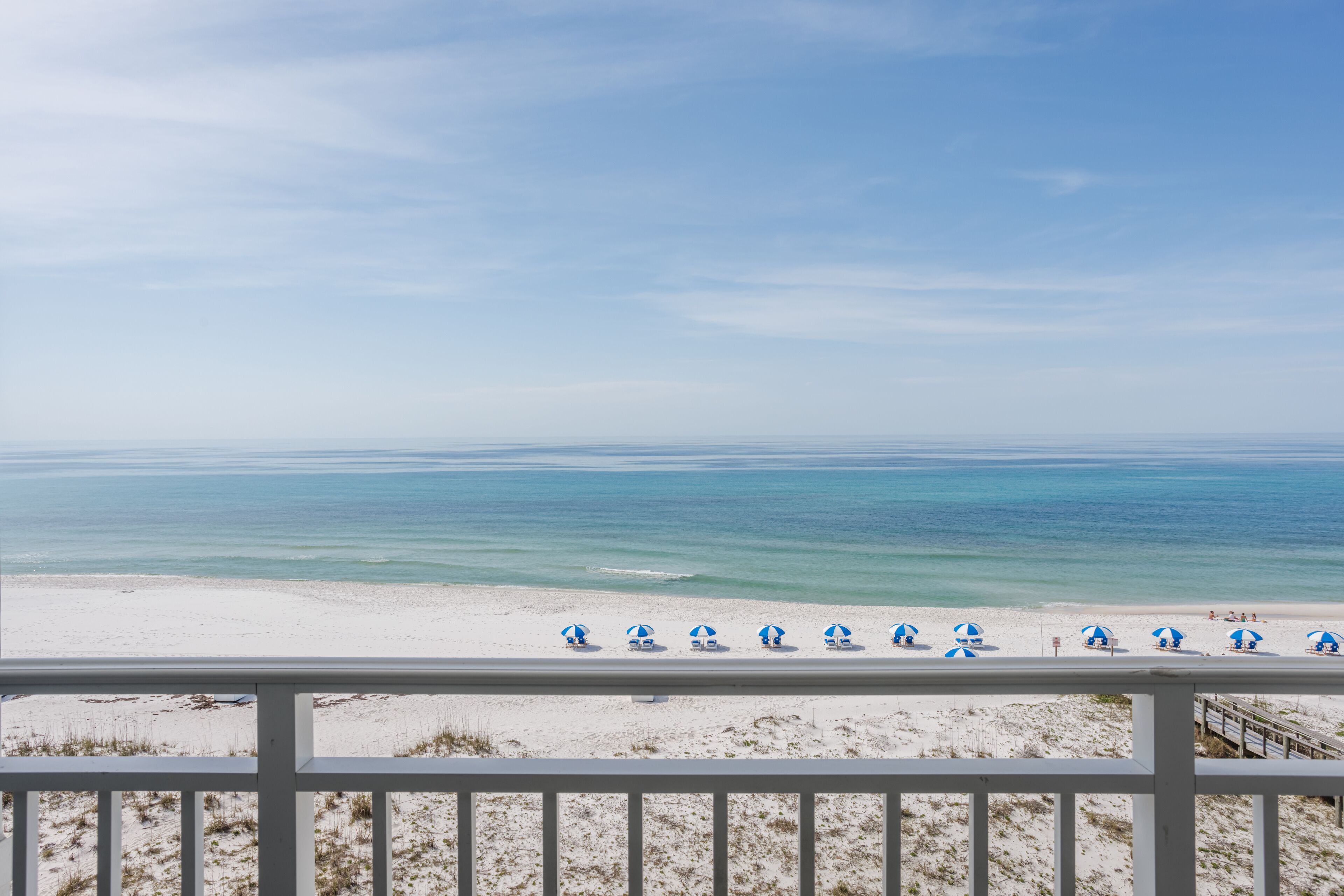 A beach view with a white railing and several blue umbrellas lined up along the shore.