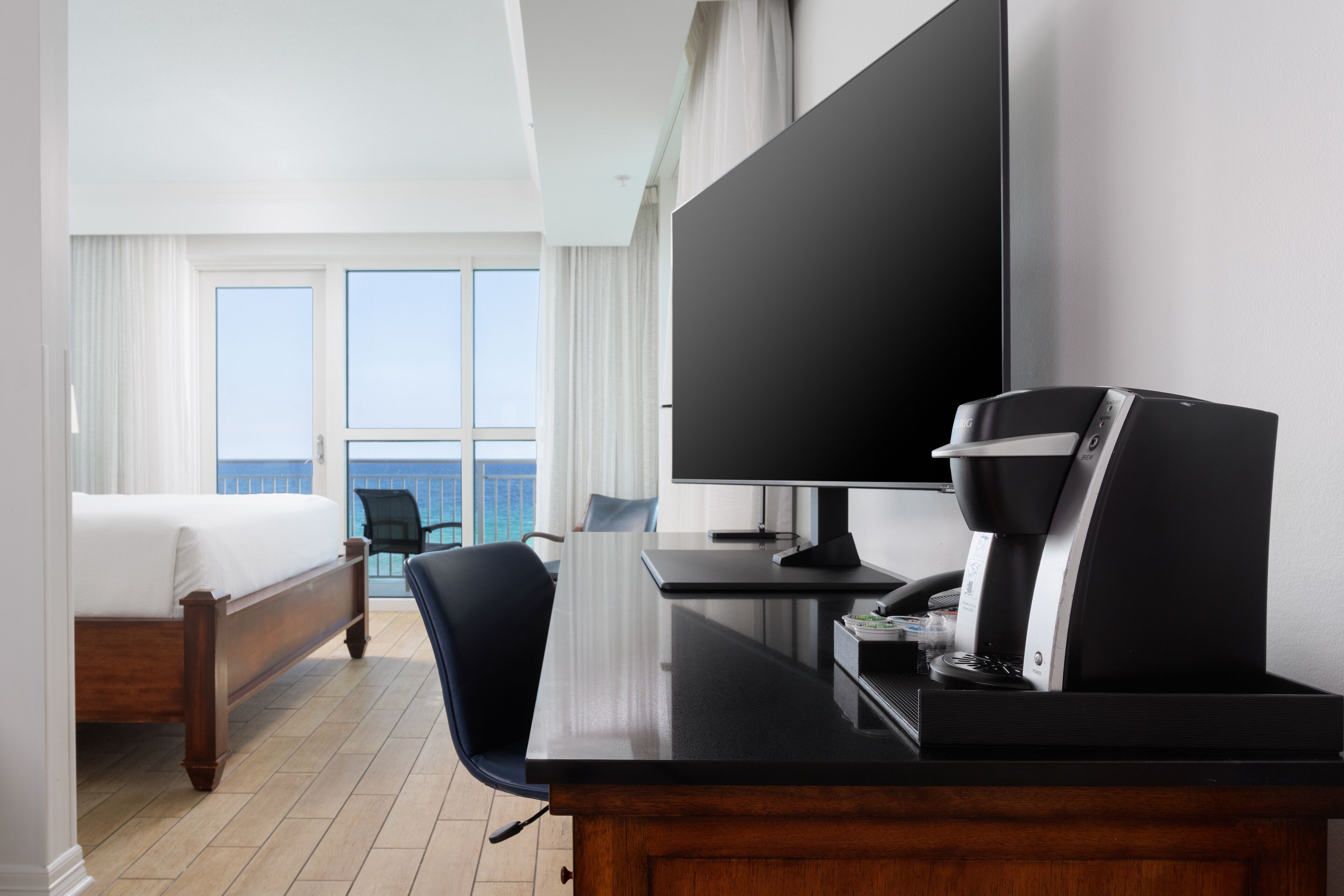 A hotel room with a bed, a desk with a coffee maker, a TV, and a view of the ocean through large windows.