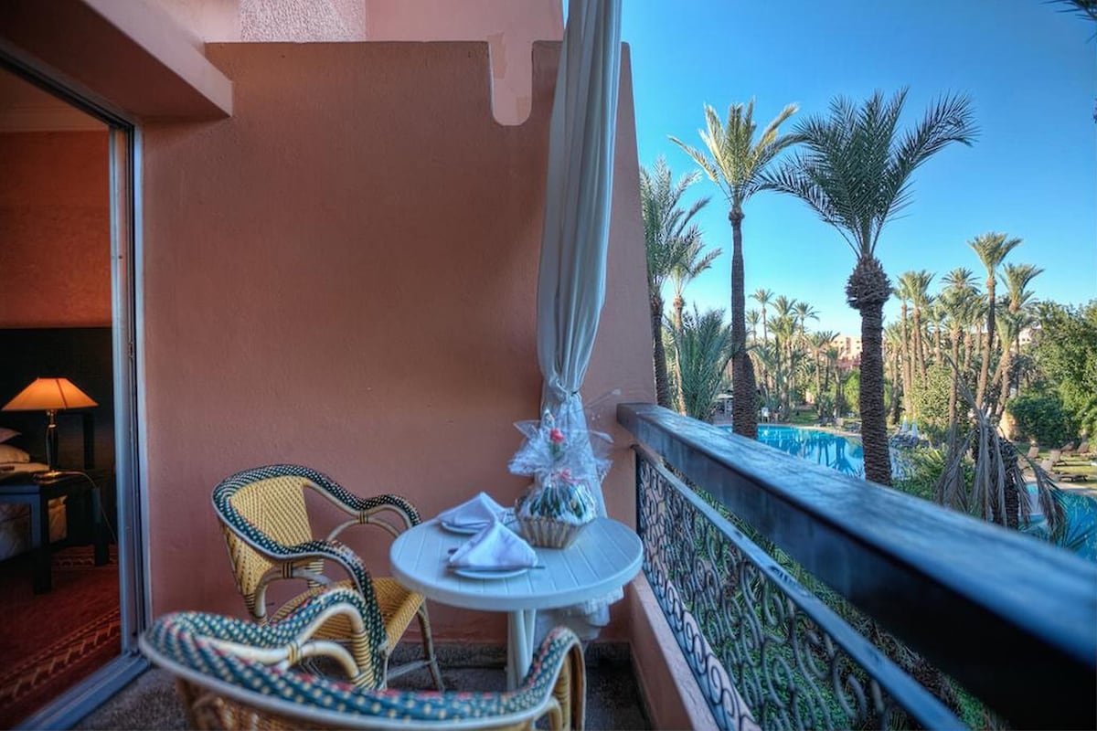A balcony with wicker chairs and a table, overlooking a pool and palm trees.
