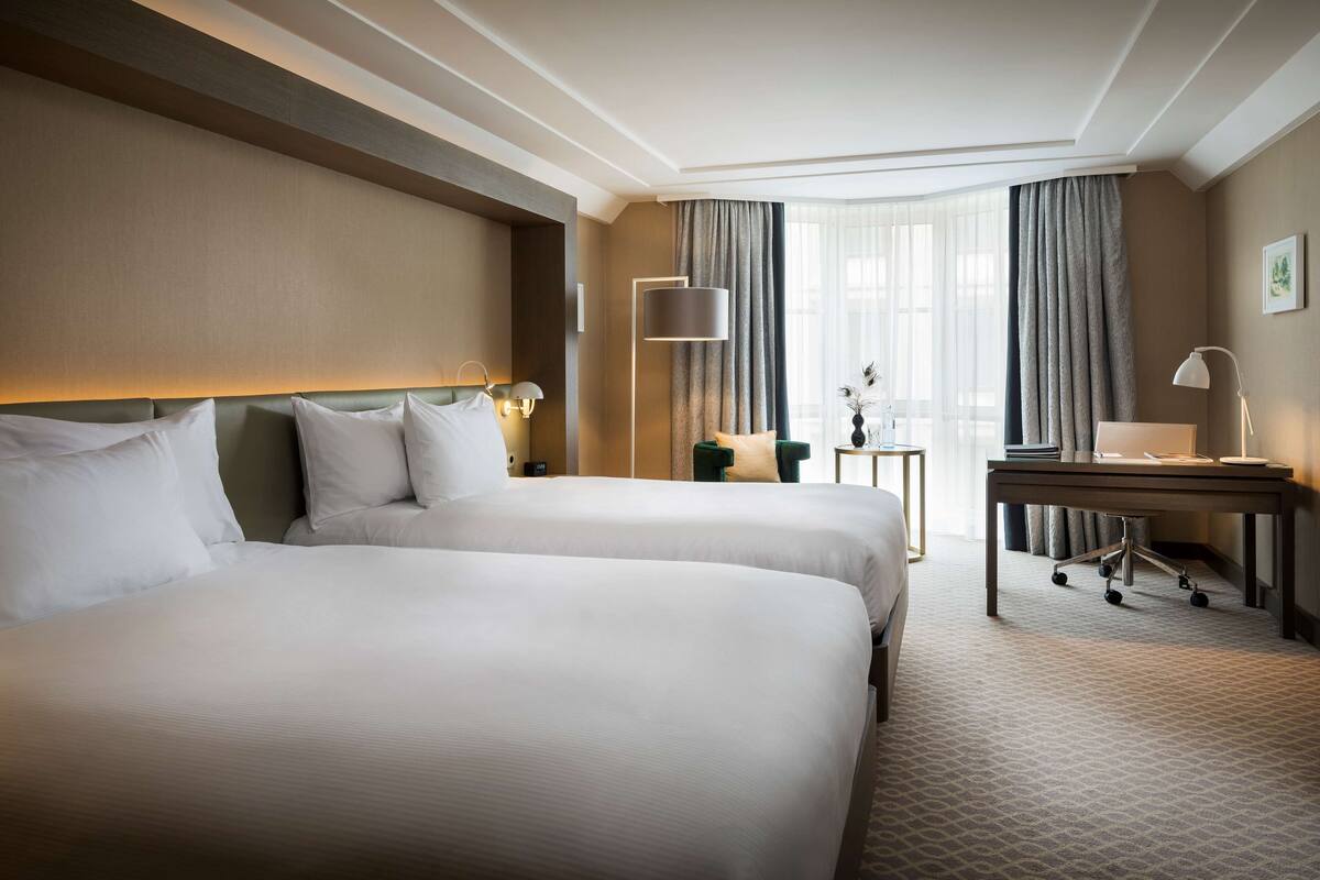 A hotel room with two beds, a desk, a lamp, and a view of the outdoors.
