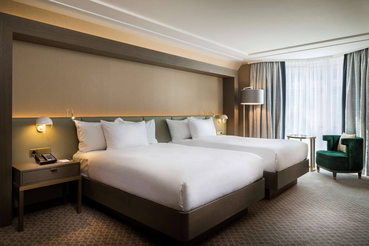 A hotel room with two beds, a nightstand with a phone, a lamp, and a chair by the window.