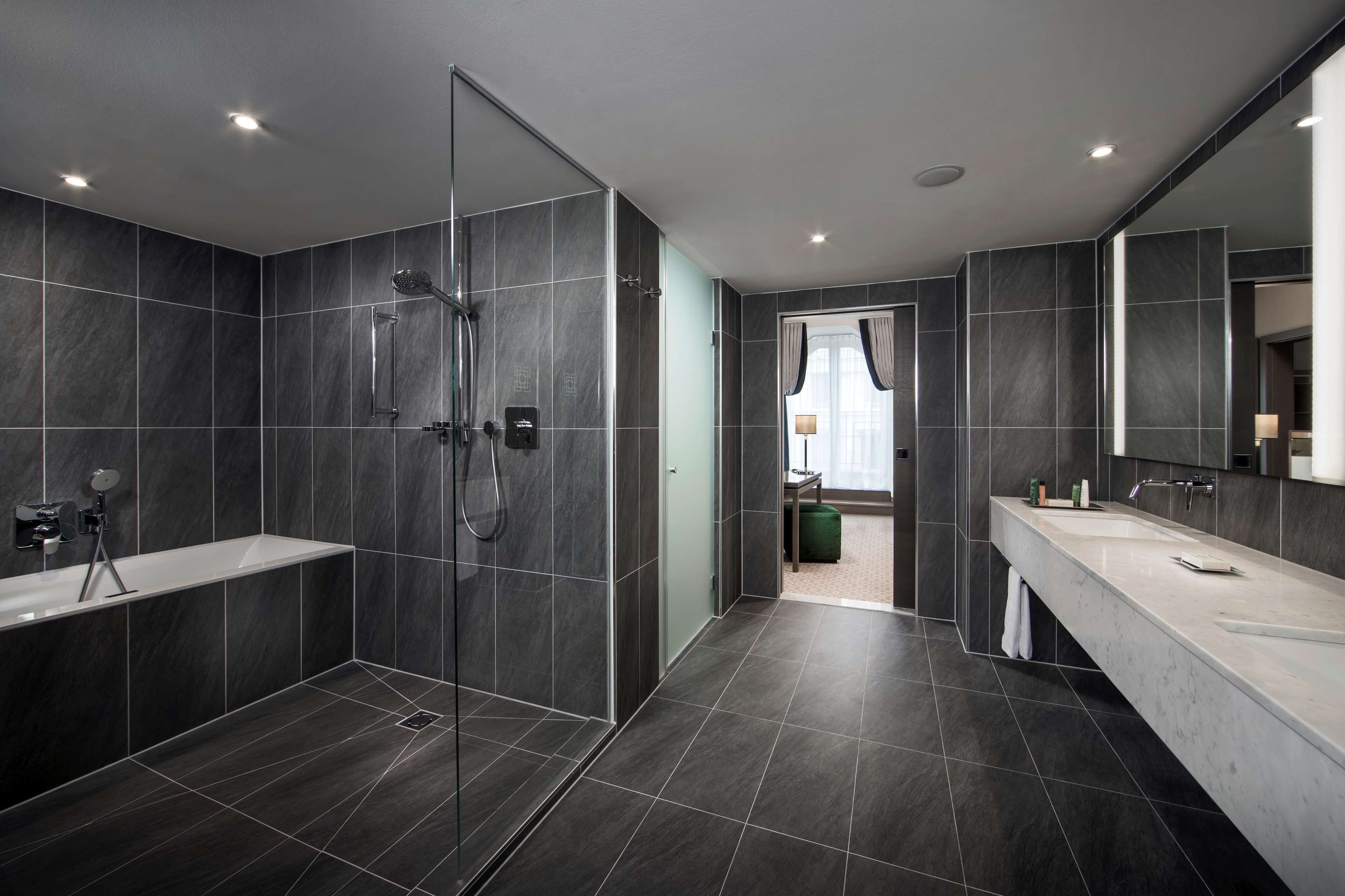 A modern bathroom with a glass-enclosed shower, a large vanity with a mirror, and dark tiled walls.