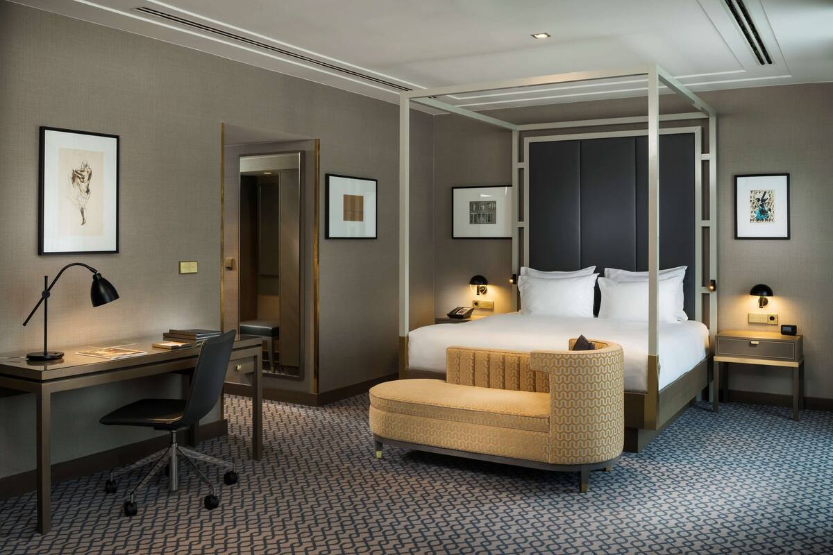 A hotel room with a four-poster bed, a desk with a chair, a bedside table with a lamp, and a small ottoman.