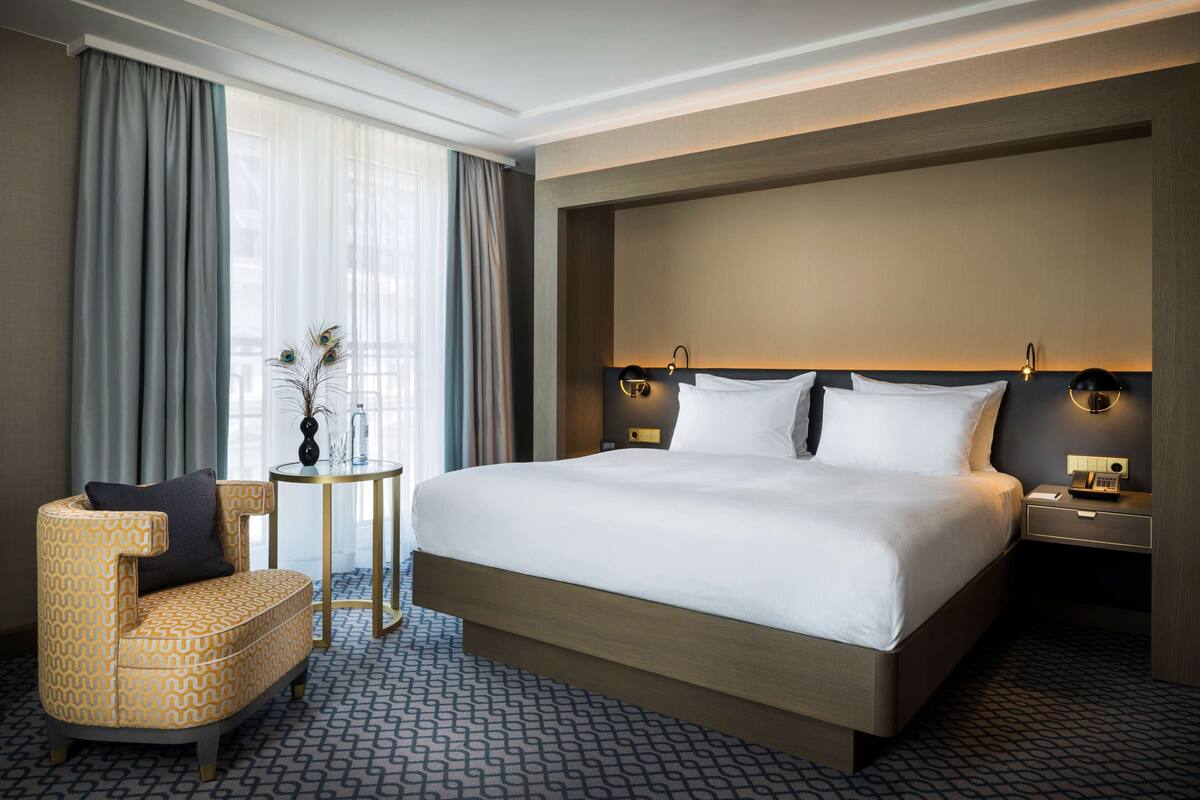 A hotel room with a large bed, a chair, a small table, and a nightstand with a lamp.