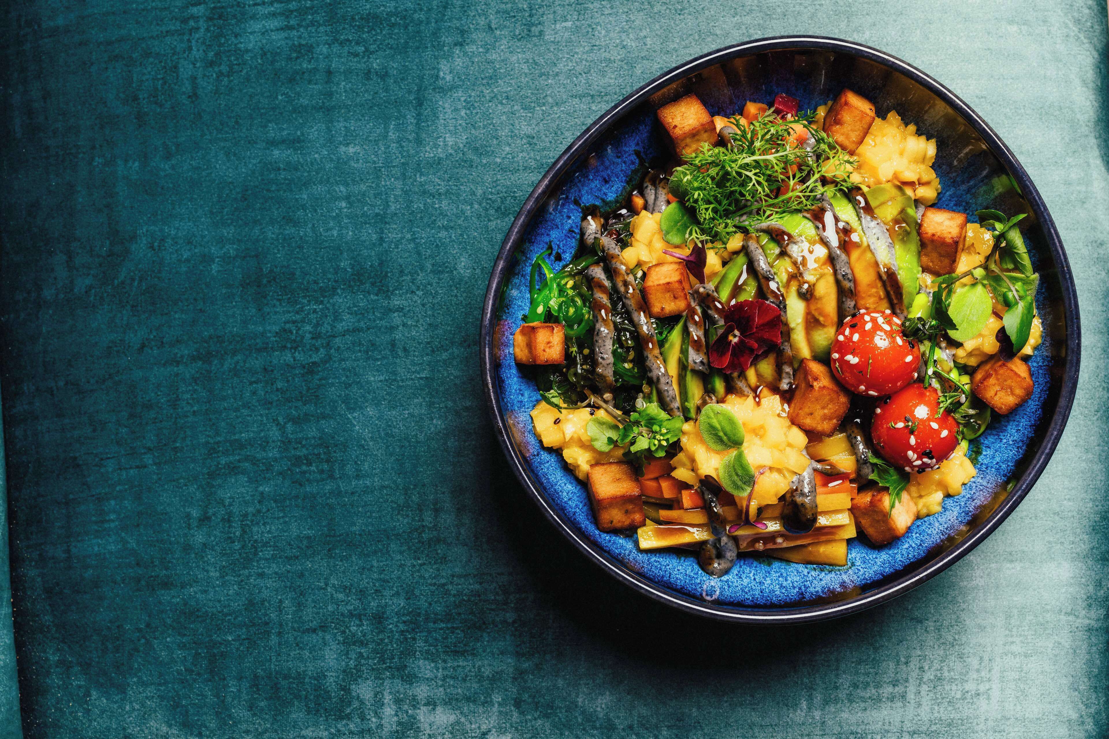 A bowl of colorful salad with various vegetables and tofu on a textured teal background.
