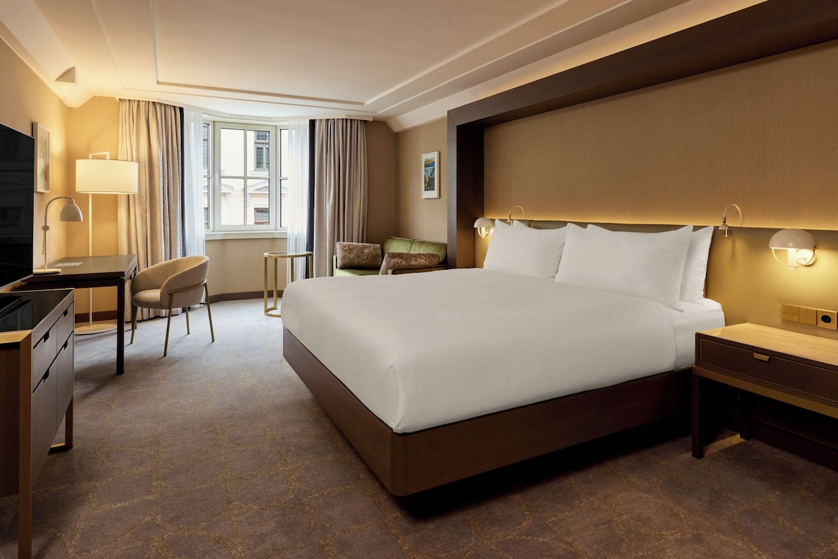 A hotel room with a large bed, a desk, a chair, a bedside table, and a lamp.
