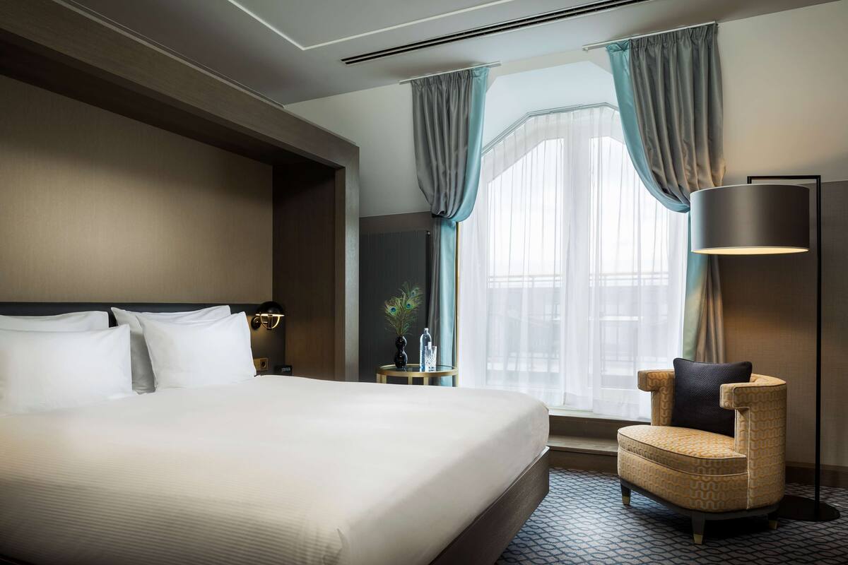 A hotel room with a large bed, a chair, a floor lamp, and a window with curtains.