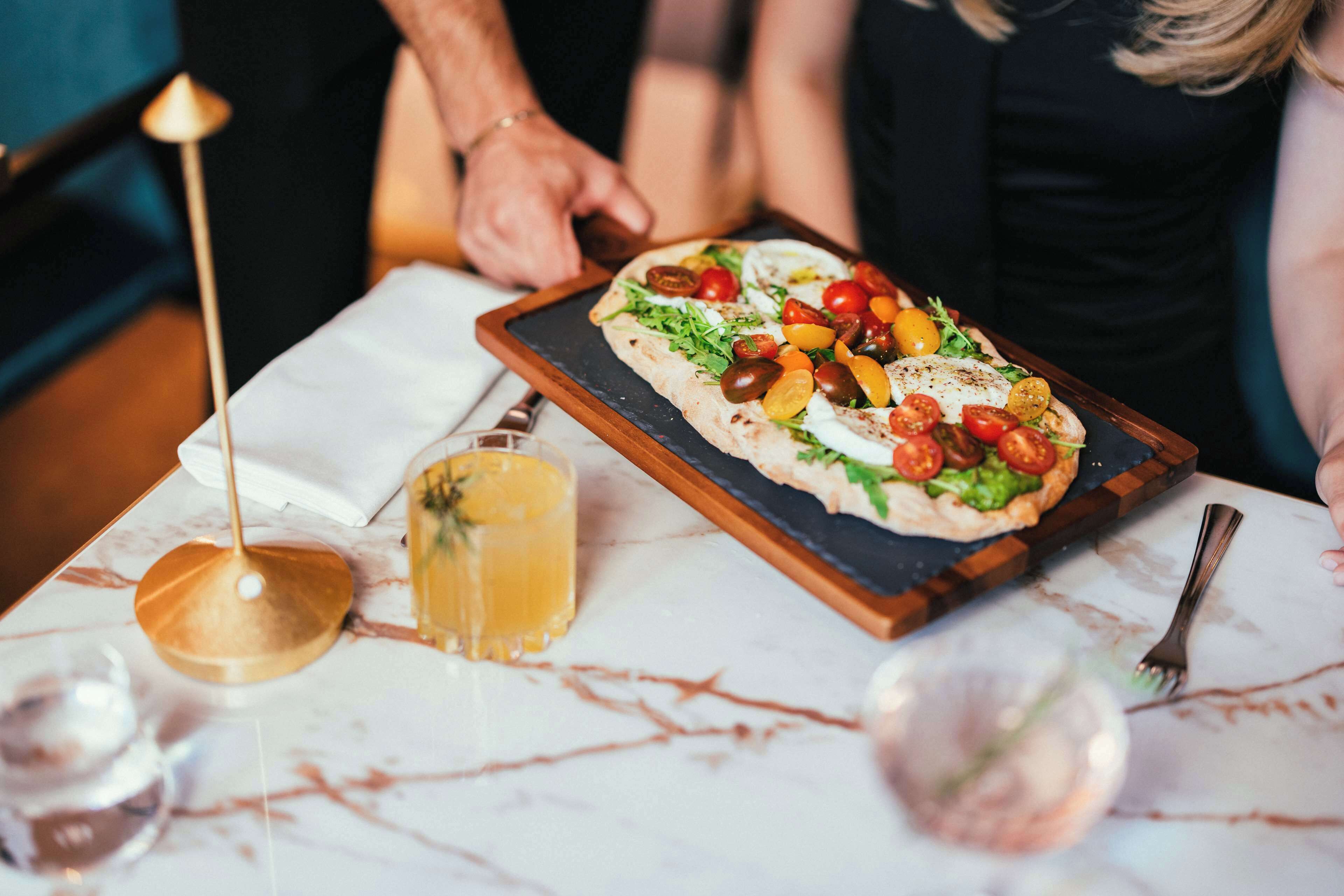 A pizza on a wooden board with a white napkin underneath, a glass of yellow drink, and a fork on a marble table.