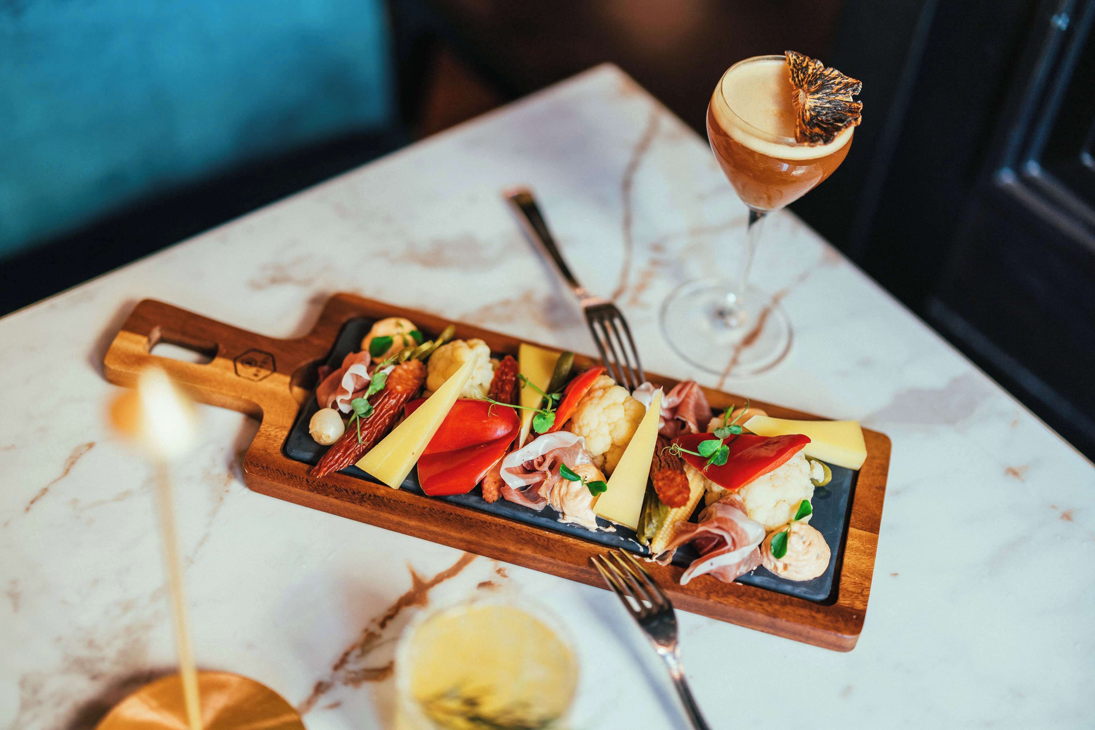 A wooden board with various cheeses, meats, and vegetables, accompanied by a glass of cocktail and a fork on a marble table.