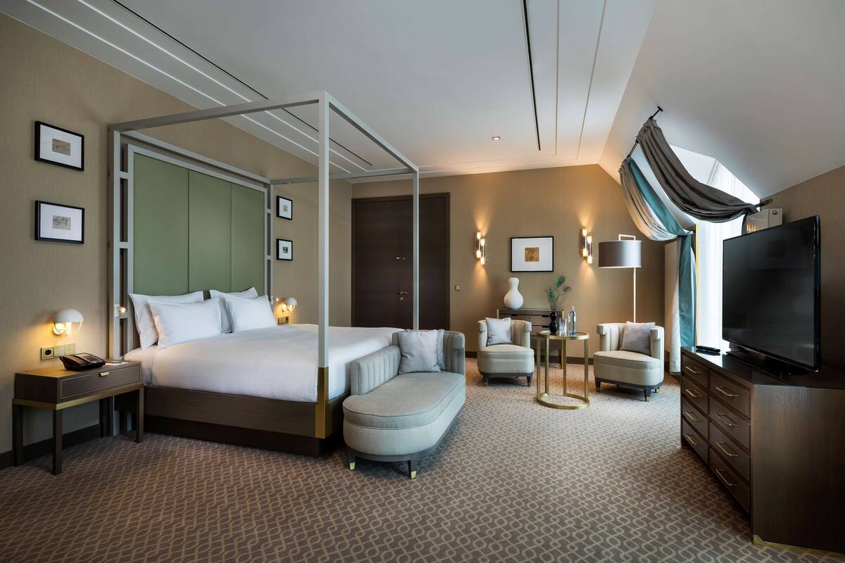 A modern hotel room with a four-poster bed, a seating area with two armchairs, a flat-screen TV, and a large window with curtains.