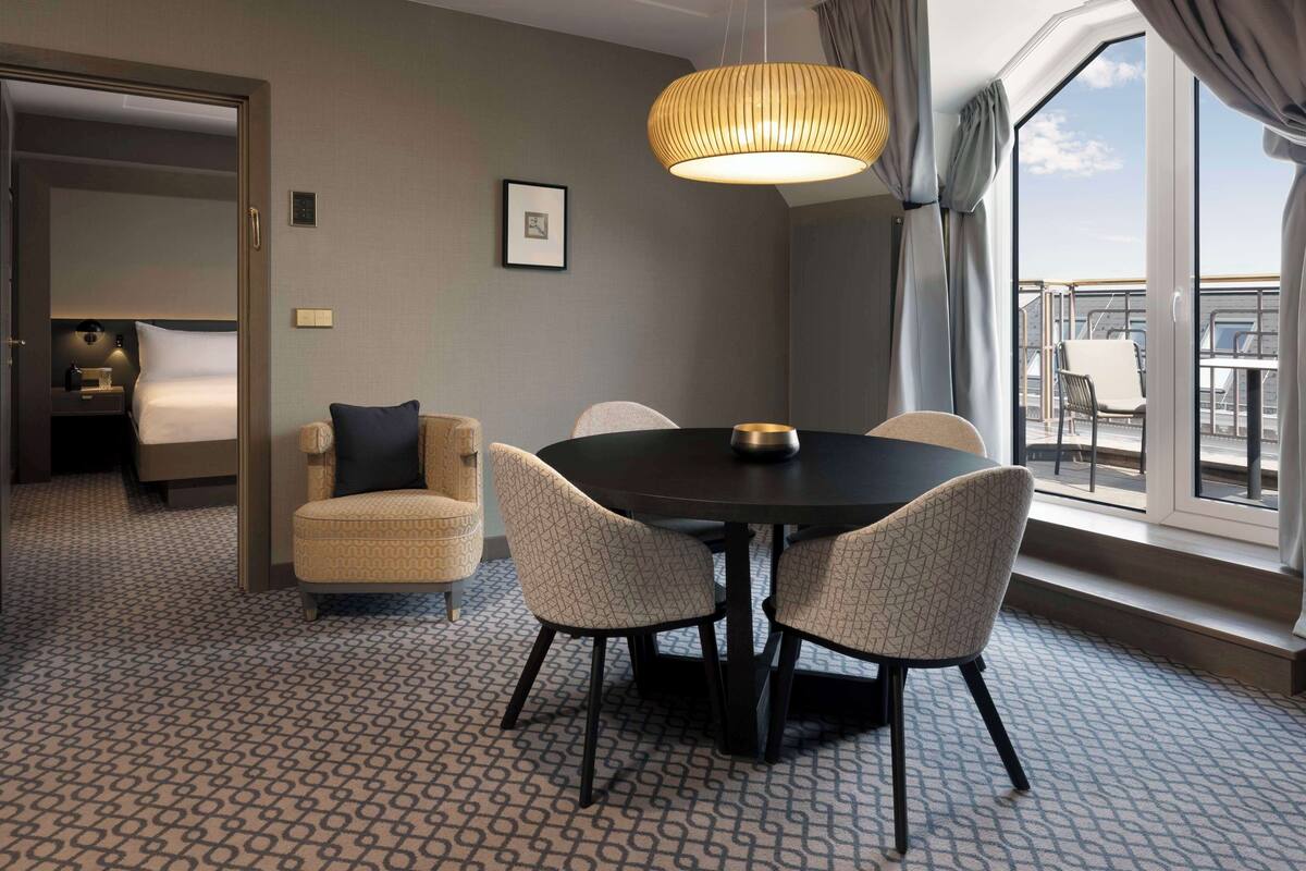 A hotel room with a dining area featuring a round table and four chairs, a large window with a view, and a patterned carpet.