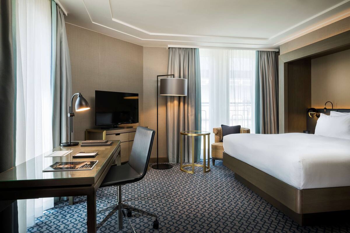 A hotel room with a bed, desk, chair, TV, lamp, and a small table.