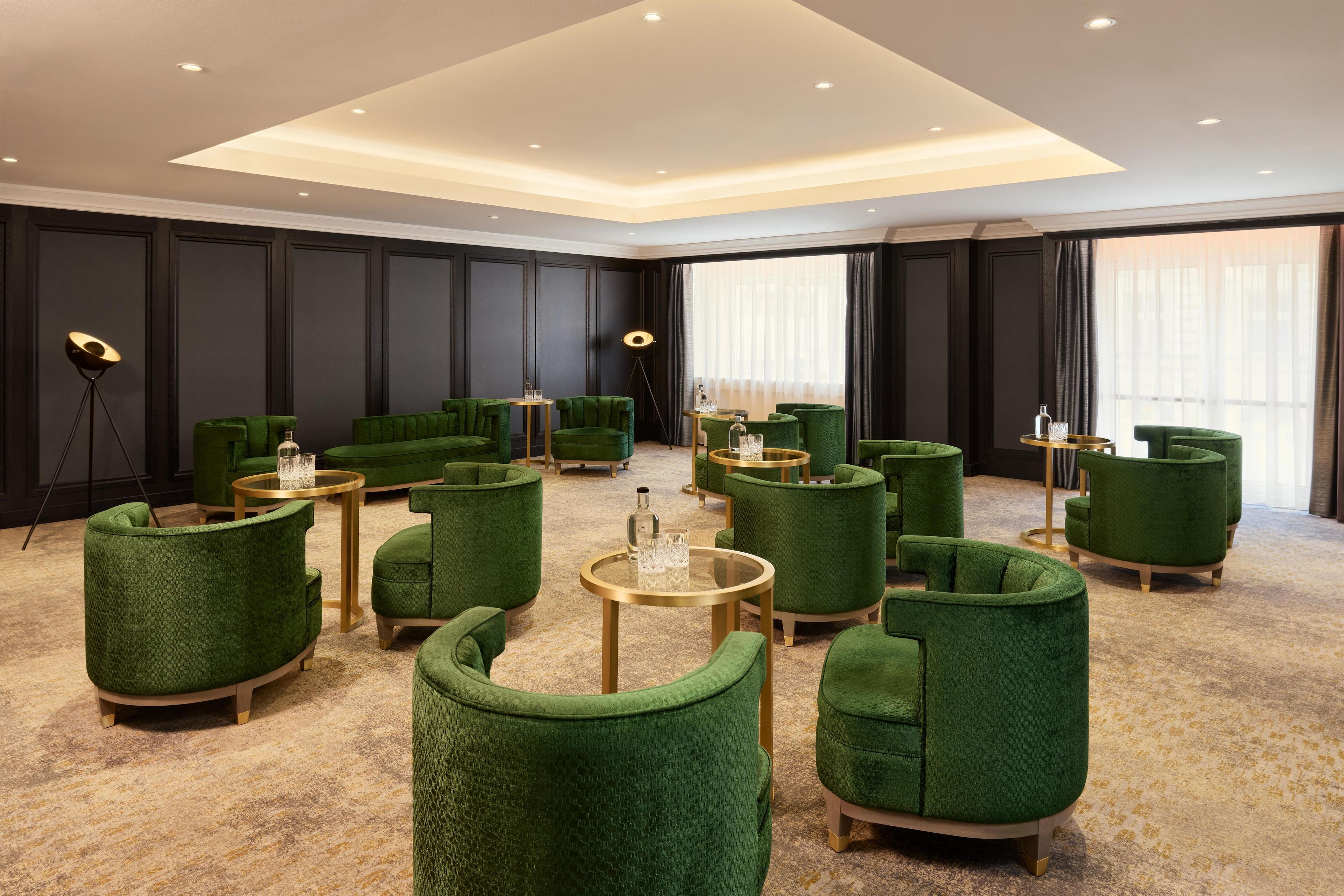 A modern lounge area with green upholstered seating and gold accents, featuring a high ceiling and large windows.