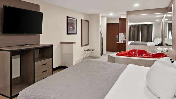 Habitación de hotel con cama, televisor montado en un mueble de madera, bañera roja y un espejo grande.