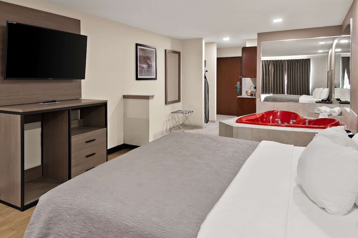 A hotel room with a bed, a TV mounted on a wooden cabinet, a red bathtub, and a large mirror.