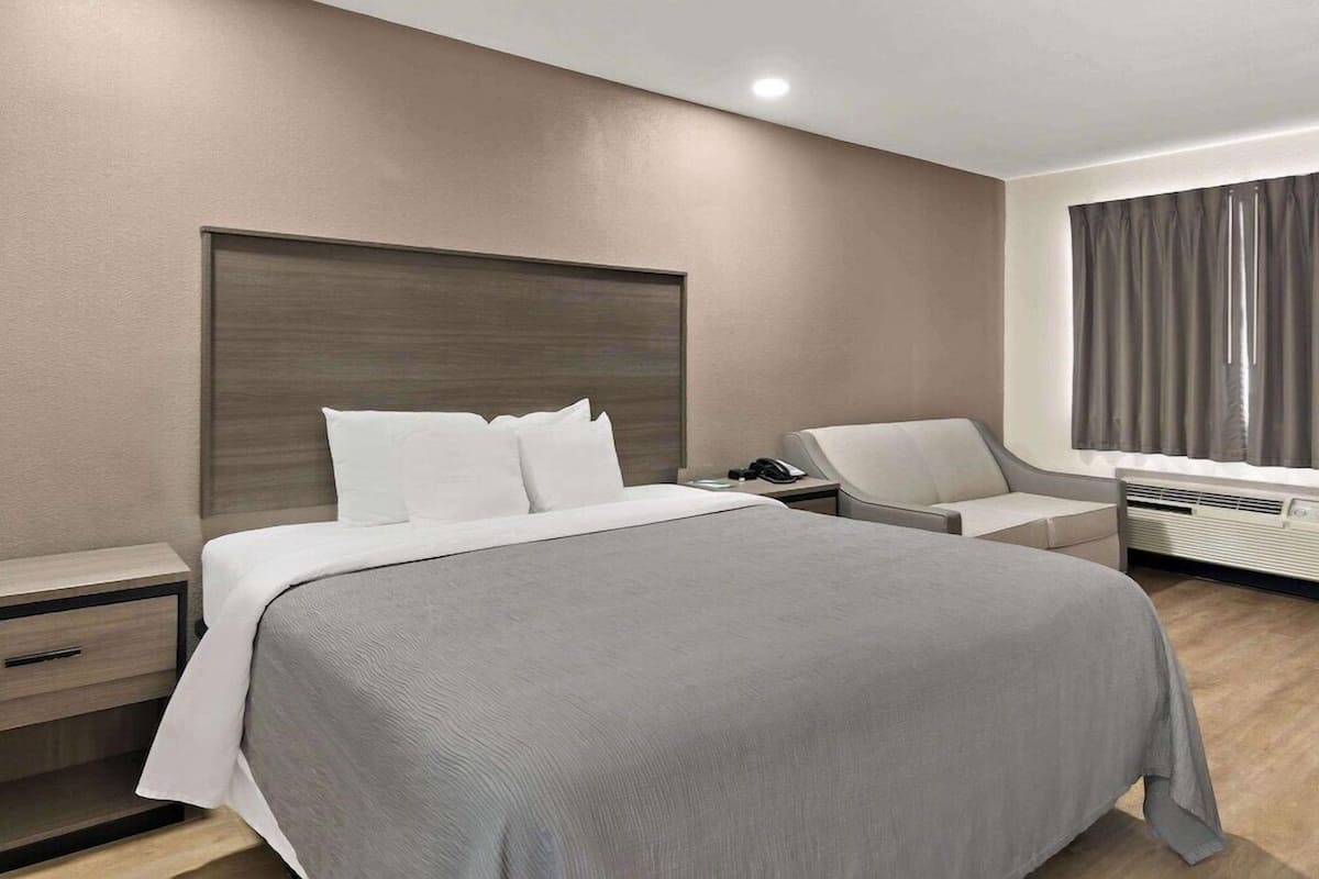 A modern hotel room with a large bed, a bedside table with a phone, a chair, and a window with curtains.