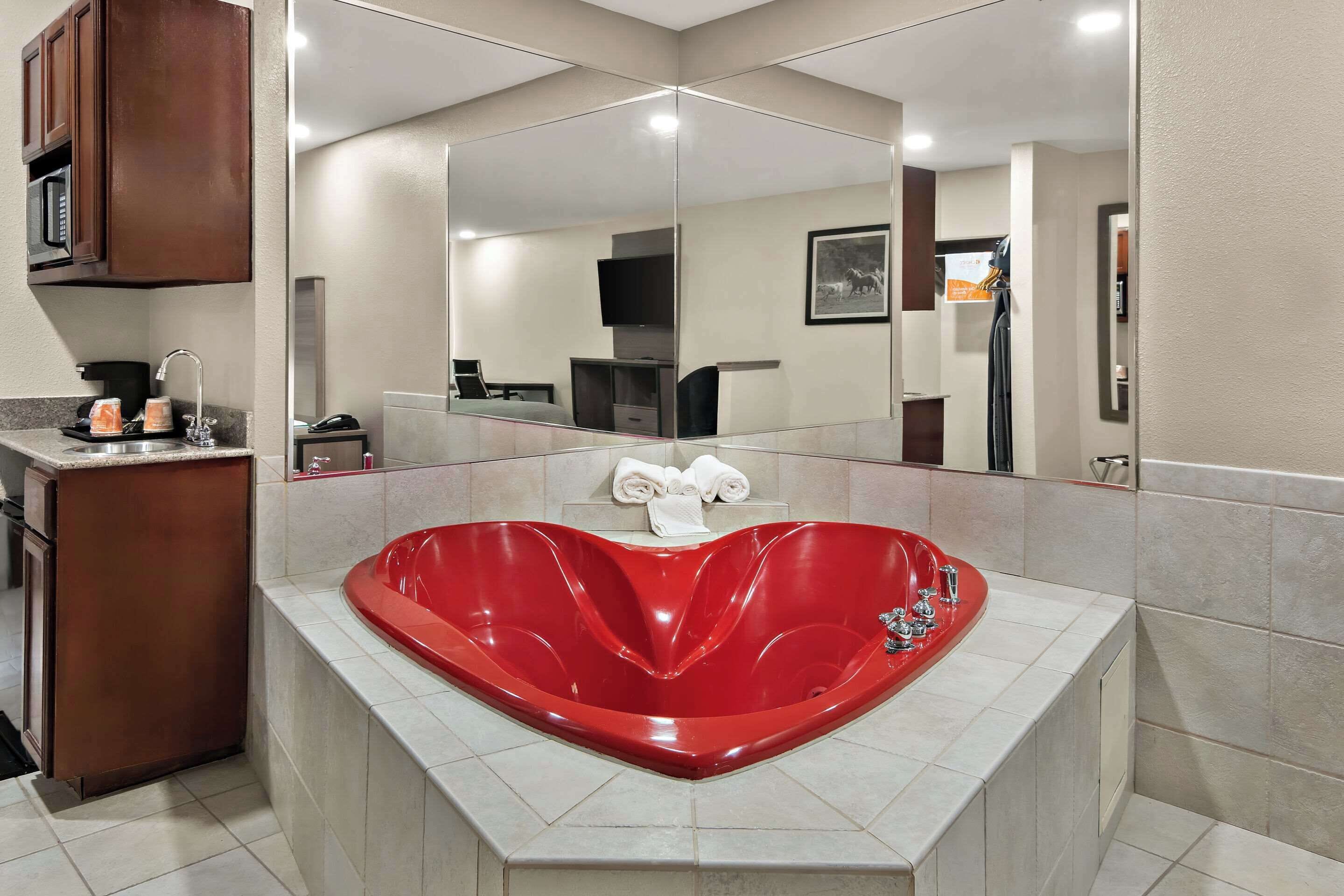 A modern bathroom with a heart-shaped red bathtub, a large mirror, and a vanity with a sink and towel rack.