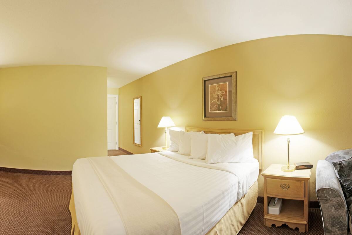 Habitación de hotel con una cama grande, dos mesitas de noche con lámparas, un pequeño sofá y una puerta que da a otra habitación.