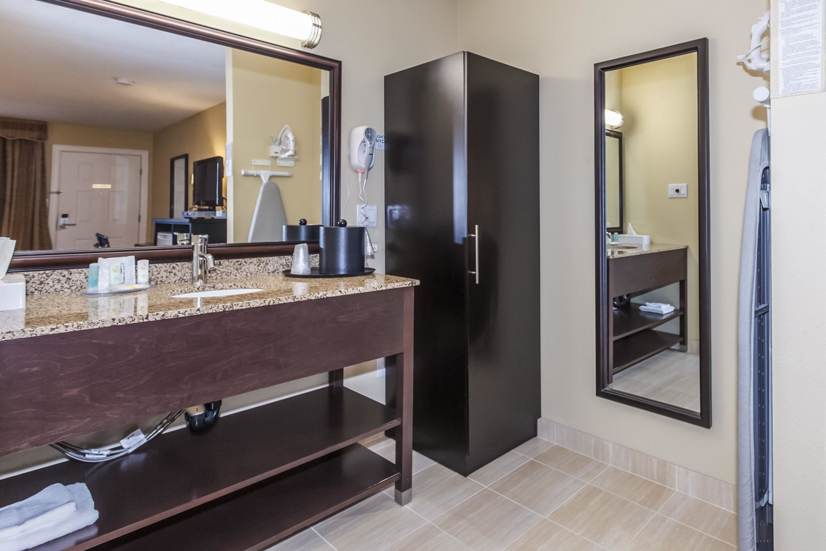 A modern bathroom with a granite countertop, dark cabinetry, a black refrigerator, and a large mirror.