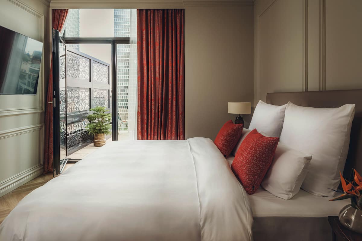 Ein Hotelzimmer mit einem Bett, einem Nachttisch, einer Lampe, einer Topfpflanze und Blick auf die Stadt durch ein Fenster.