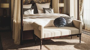 A four-poster canopy bed with a chandelier, a patterned carpet, and a nightstand with a lamp.