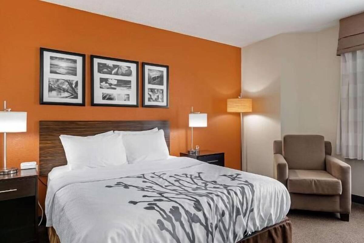 Quarto de hotel com uma cama, duas mesas de cabeceira, uma cadeira, uma janela com cortinas e três quadros na parede.