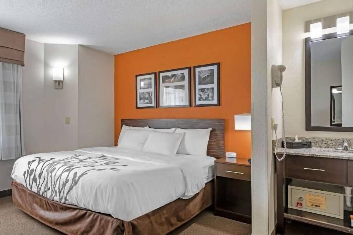 Quarto de hotel com cama, escrivaninha, cadeira, janela com cortinas, parede com detalhes em laranja e quadros.