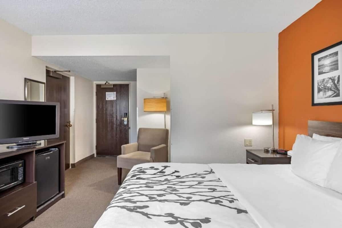 Quarto de hotel com cama, televisão, cadeira, mesa de cabeceira e porta que dá para outro aposento.