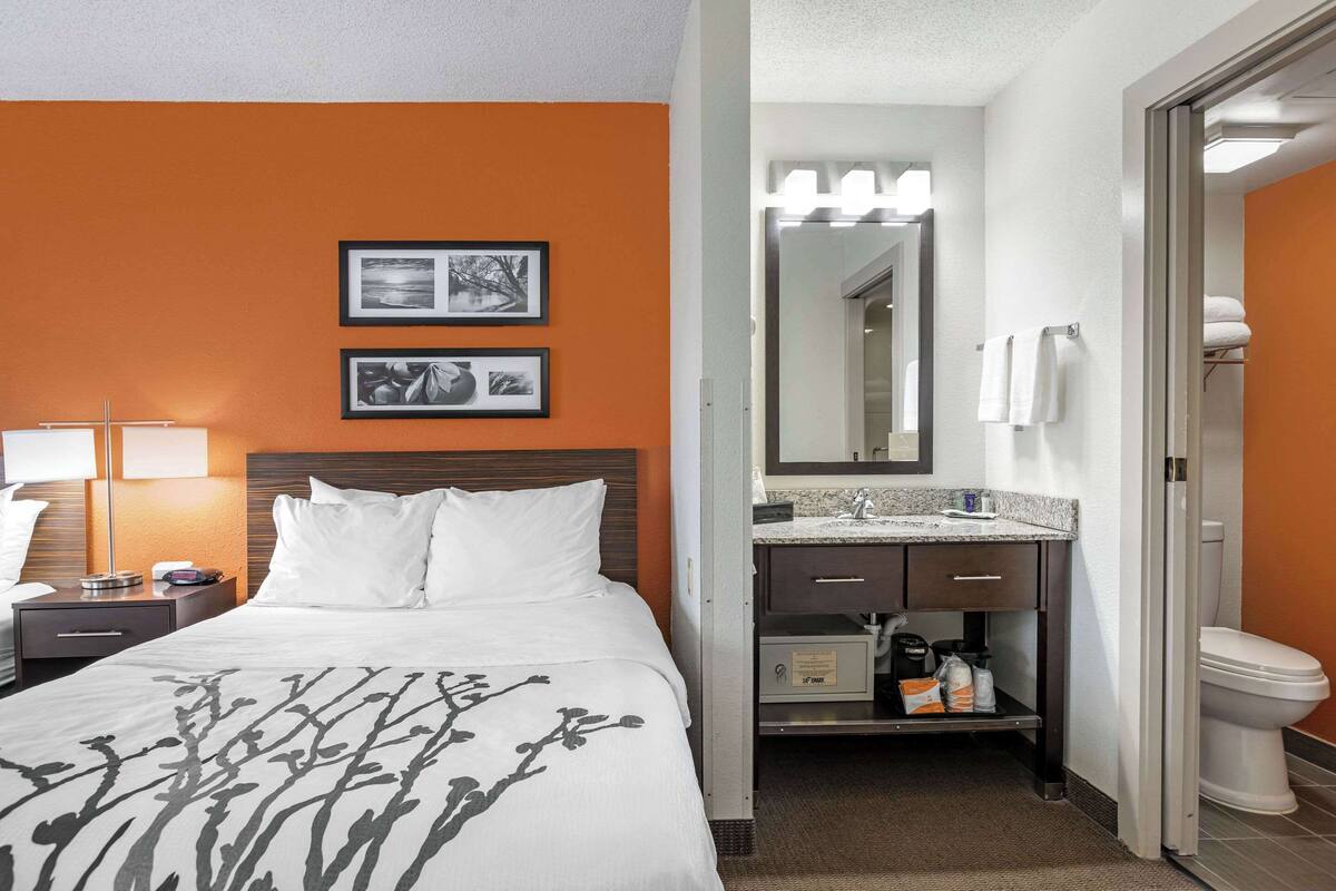 Quarto de hotel com cama, mesa de cabeceira, luminária e banheiro com pia, espelho e vaso sanitário.