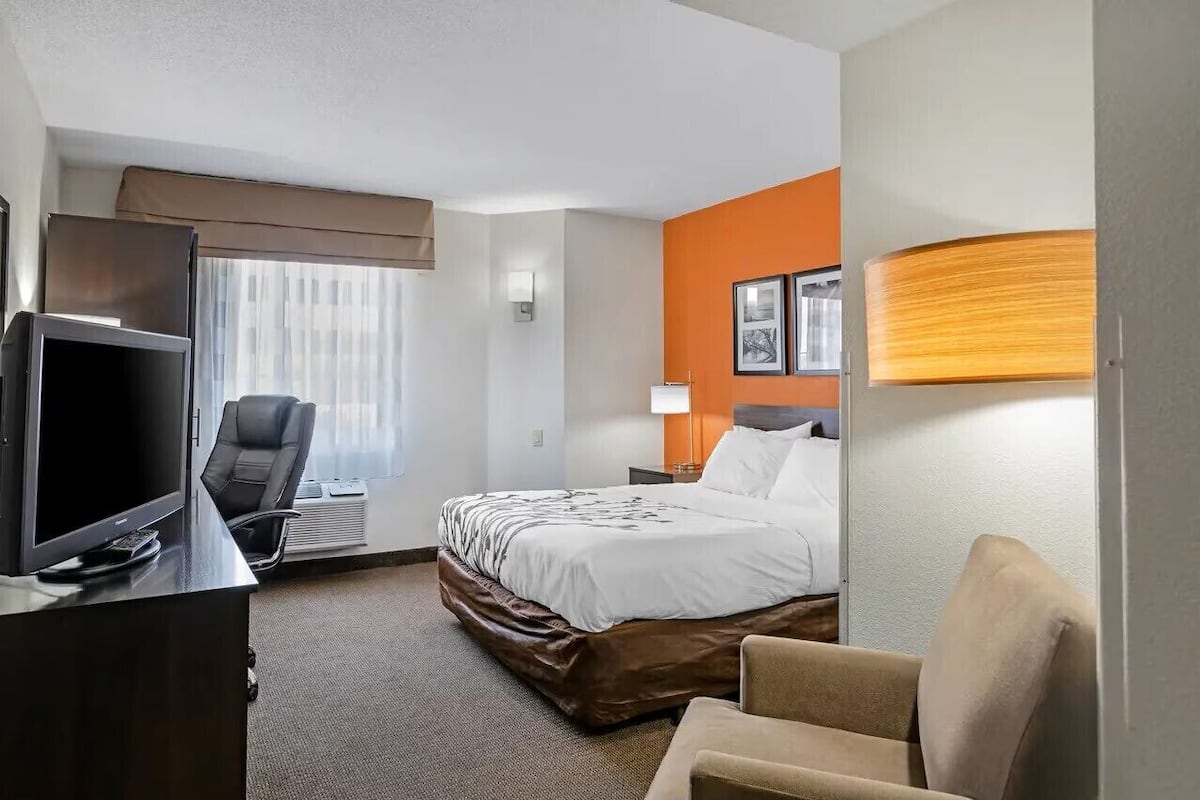 Quarto de hotel com cama, escrivaninha com televisão, cadeira, abajur e janela com cortinas.